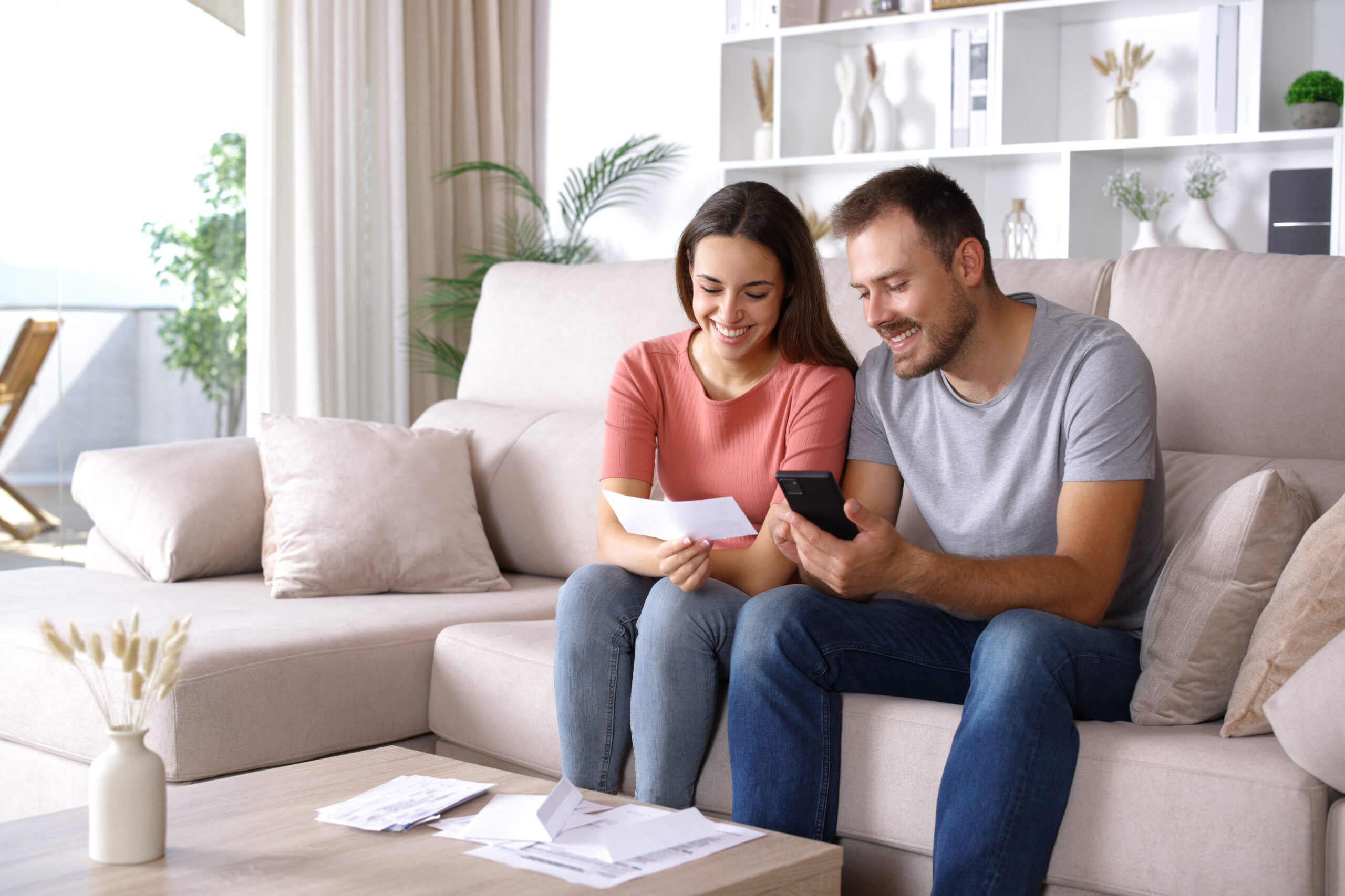 Happy couple checking bank statement and smart phone sitting on a couch at home