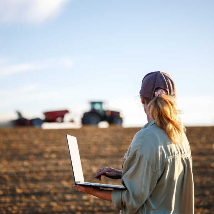 Smart farming. Woman farmer with laptop analyzing agricultural data in front of tractor and seeding machine on cultivated field