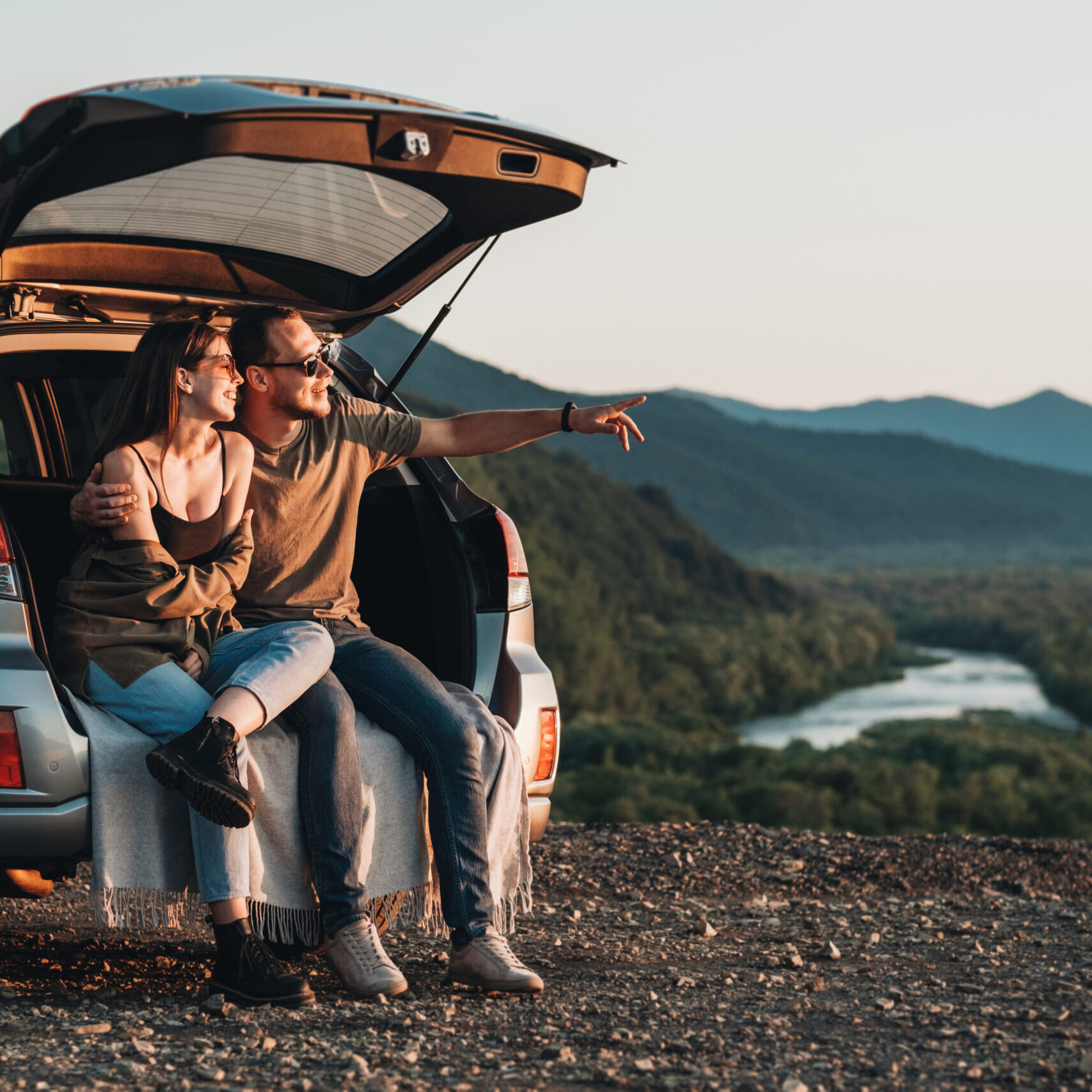 Young Traveler Couple on Road Trip, Man and Woman Sitting on the Opened Trunk of Their Car Over Sunset