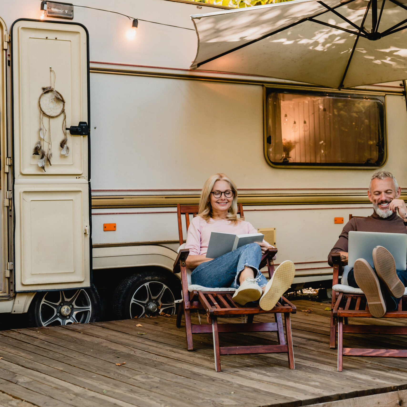 Senior joyful couple relaxing in the porch of thier motorhome with book and laptop