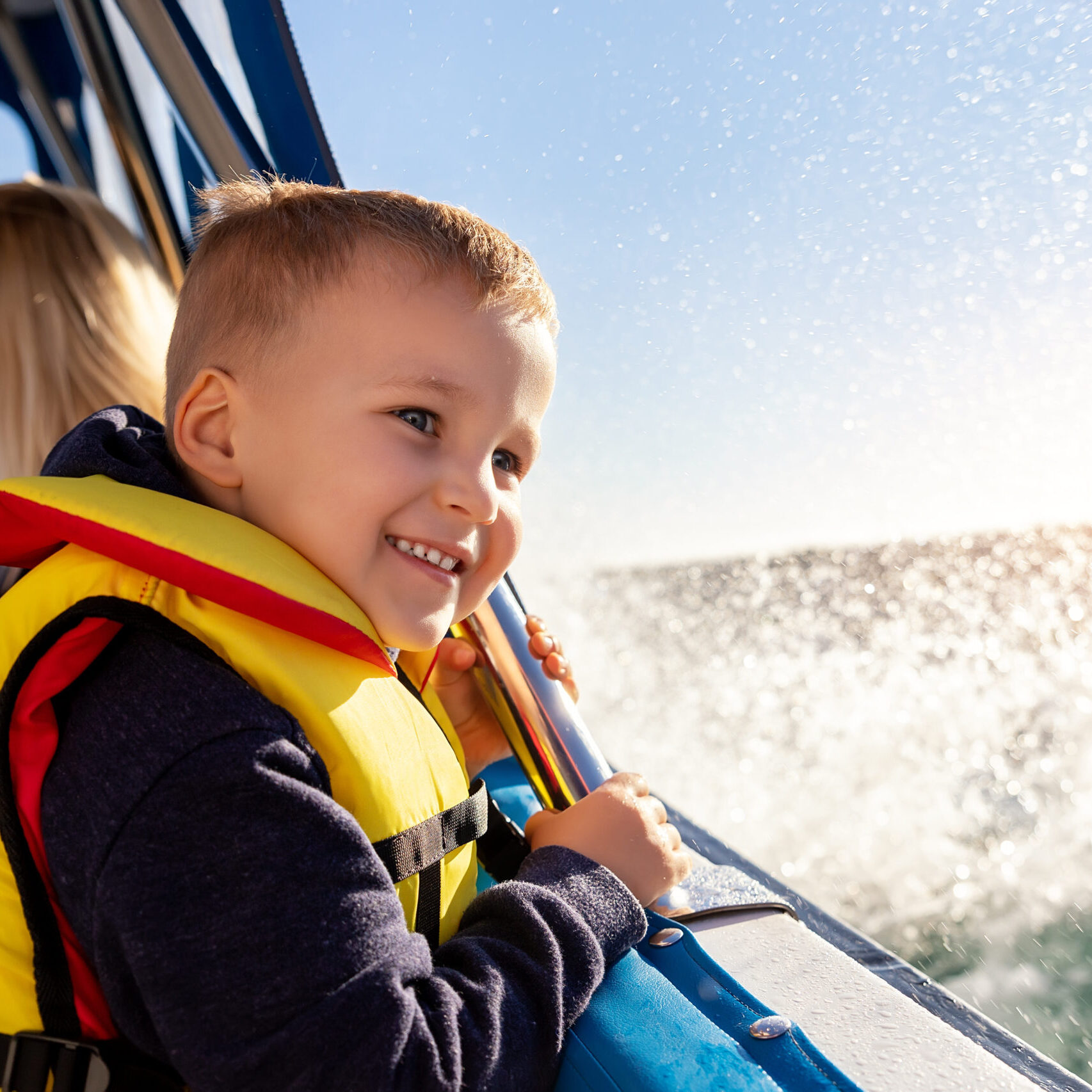 Portrait of cite little blond happy excited smiling caucasian boy wear lifevest enjoy sailing on motor boat sea against blue sky and water splash wave sun backlit. Summer travel vacation recreation.
