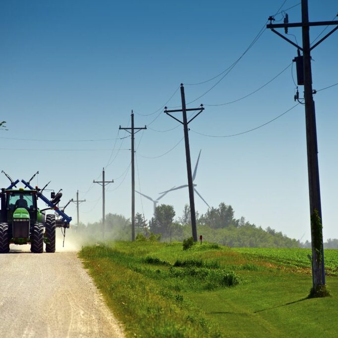Minnesota Farmer on Suburb Road Riding Tractor with Farm Equipment. Old Wood Electric Poles and Wind Turbines in a Background. Minnesota, USA.